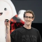 Person smiling, holding a small black device on stage, wearing a "Building is my love" shirt, large plush mascot displayed...