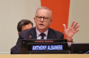 Man speaking at a conference, gesturing with one hand. Nameplate reads Anthony Albanese. Orange and beige background.