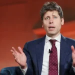 A man in a suit with a polka dot tie gestures while speaking, seated against an orange background.