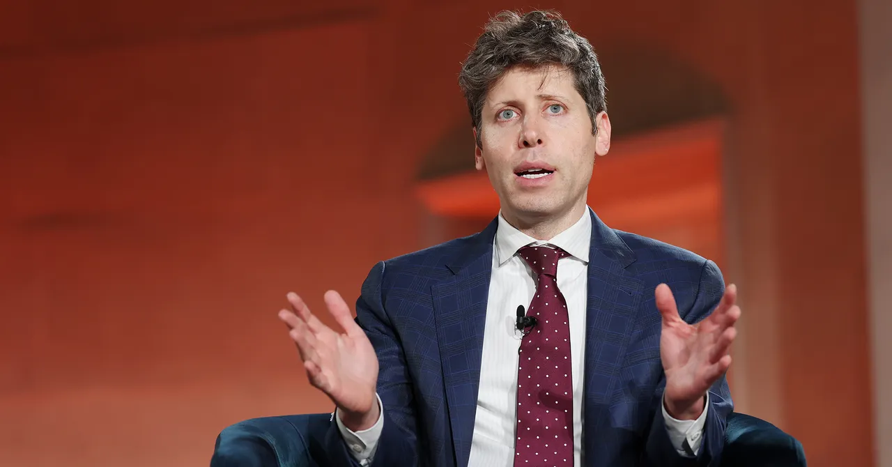 A man in a suit with a polka dot tie gestures while speaking, seated against an orange background.