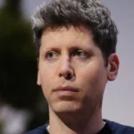 A person with short hair looks to the side against a blurred background, wearing a dark shirt.
