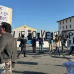 Protesters holding signs reading "People Over AI" and "No Killer Robots" gather in a street, with buildings in the backgro...
