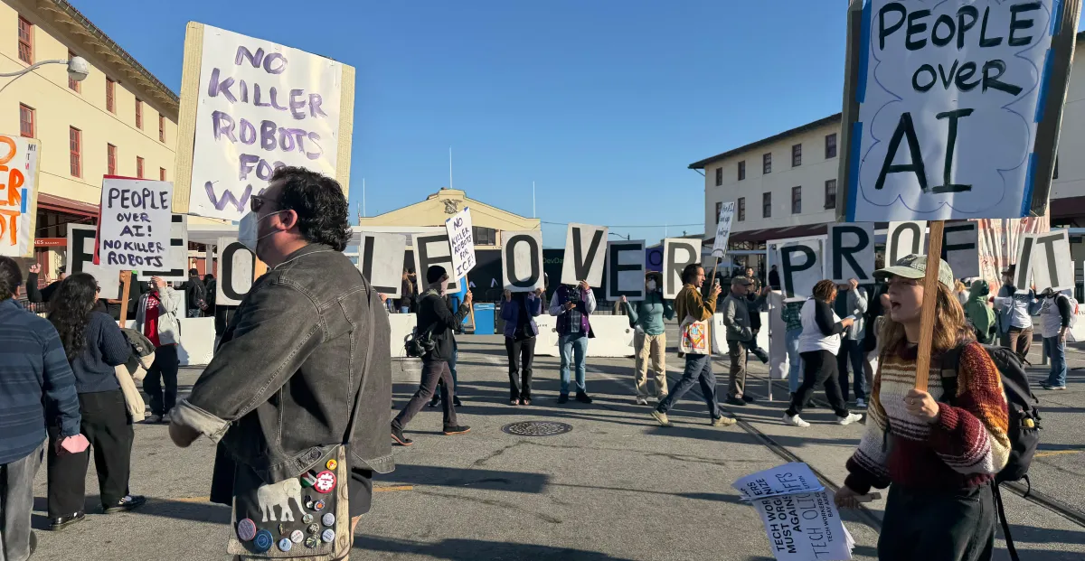 Protesters holding signs reading "People Over AI" and "No Killer Robots" gather in a street, with buildings in the backgro...