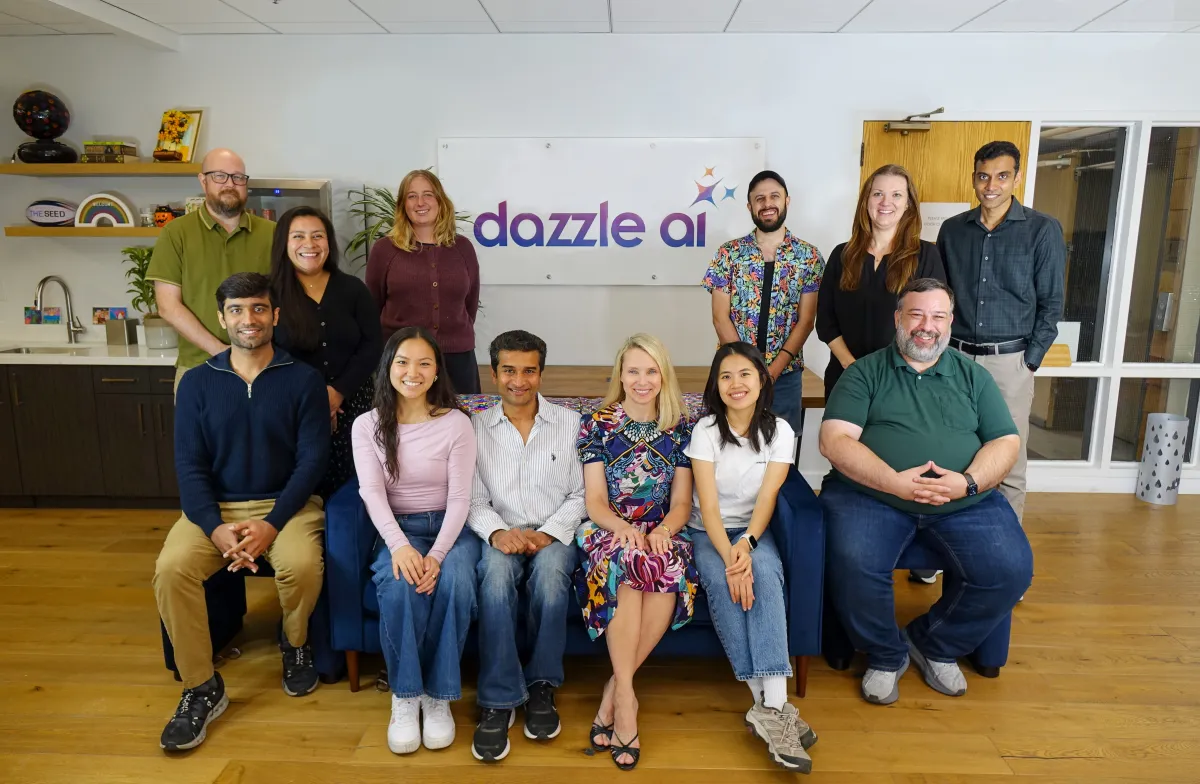 Group of people posing in an office, sitting and standing, with a "dazzle ai" sign on the wall behind them.