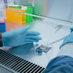 Gloved hands handling a pipette over a petri dish in a lab with colorful test tube racks in the background.