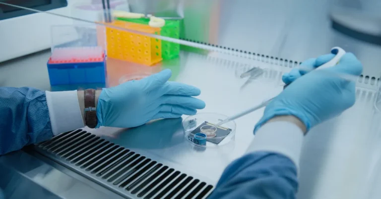 Gloved hands handling a pipette over a petri dish in a lab with colorful test tube racks in the background.