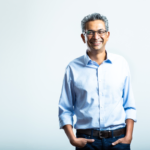 Smiling man in a light blue shirt and jeans stands against a plain white background, hands in pockets.