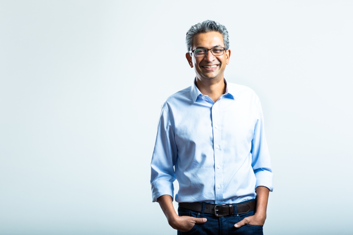 Smiling man in a light blue shirt and jeans stands against a plain white background, hands in pockets.