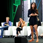 Woman presenting on stage with three seated judges listening, backdrop displays TechCrunch branding.