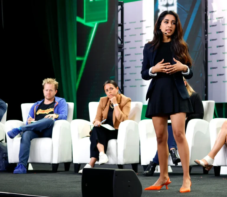 Woman presenting on stage with three seated judges listening, backdrop displays TechCrunch branding.