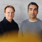 Two men stand against a textured gray wall, one in a black jacket and the other in a gray sweater, with a yellow blur in t...