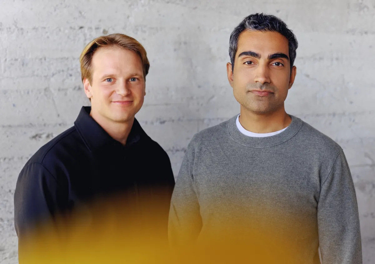Two men stand against a textured gray wall, one in a black jacket and the other in a gray sweater, with a yellow blur in t...