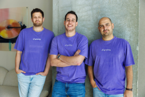 Three men in purple "Cymphony" shirts stand smiling against a concrete wall, with artwork in the background.
