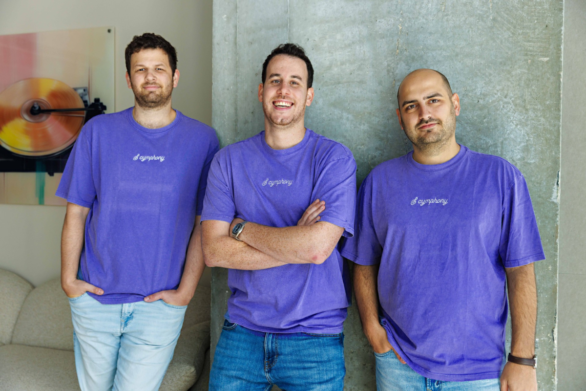 Three men in purple "Cymphony" shirts stand smiling against a concrete wall, with artwork in the background.