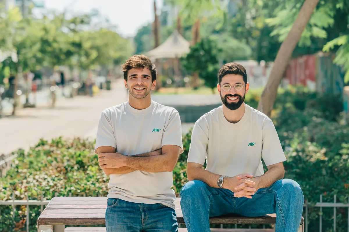 Two men in AIR-branded shirts sitting on a bench outdoors, smiling, with trees and a path in the background.