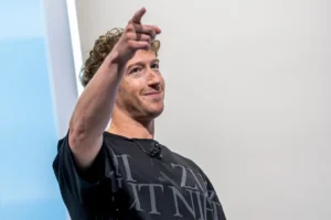 Smiling person with curly hair points forward, wearing a black shirt with white text, standing against a light background.