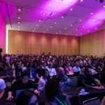 Large audience seated in a conference room with pink lighting, many using laptops, attending a tech event or seminar.