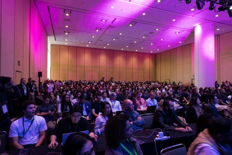 Large audience seated in a conference room with pink lighting, many using laptops, attending a tech event or seminar.
