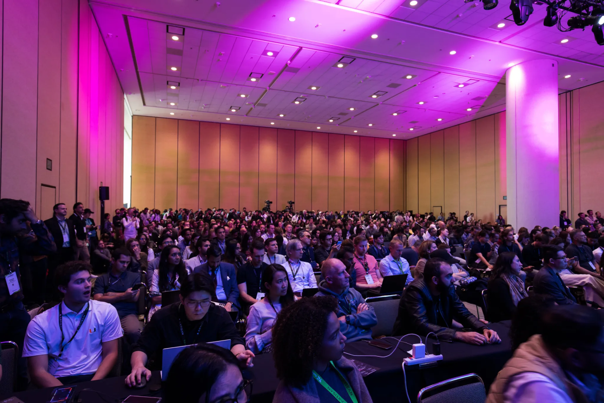 Large audience seated in a conference room with pink lighting, many using laptops, attending a tech event or seminar.