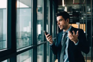 Man in suit using smartphone, gesturing with hand, standing by large office windows.