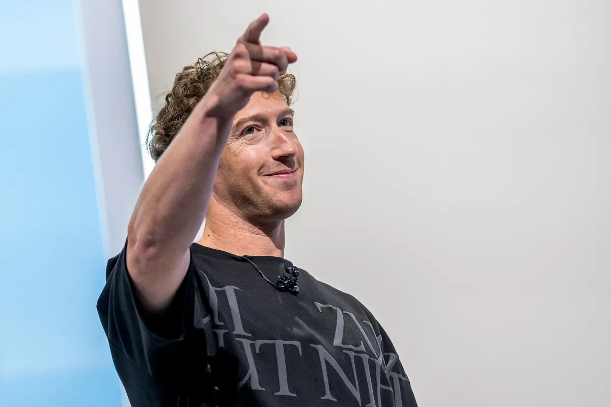 Smiling person with curly hair points forward, wearing a black shirt with white text, standing against a light background.