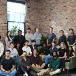 Group of diverse individuals posing in a brick-walled room with plants, some seated and others standing.