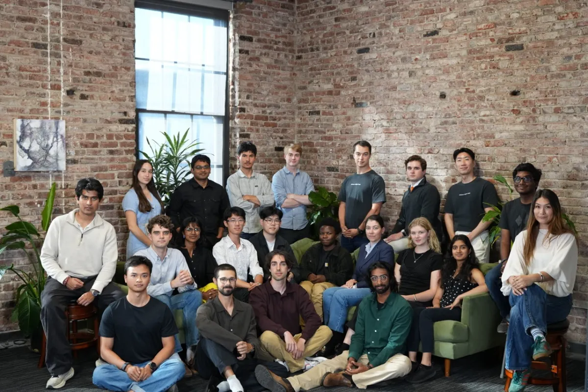 Group of diverse individuals posing in a brick-walled room with plants, some seated and others standing.