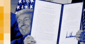 Man in blue suit holding up a signed executive order document with visible text, U.S. flag in the background.