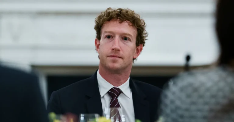 Person in a suit and tie seated at a table with blurred figures in the foreground, indoor setting.