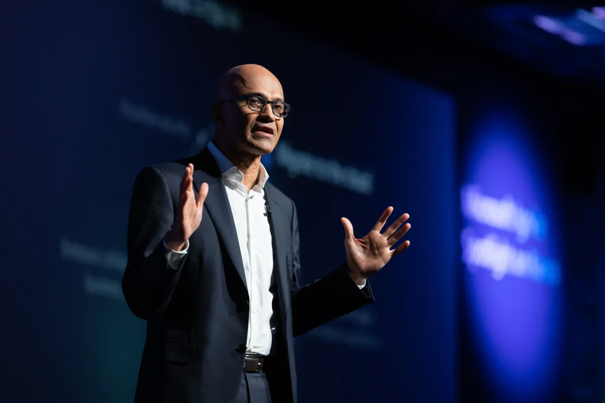 Man in a suit gestures during a presentation on a dark stage, blurred text in the background.