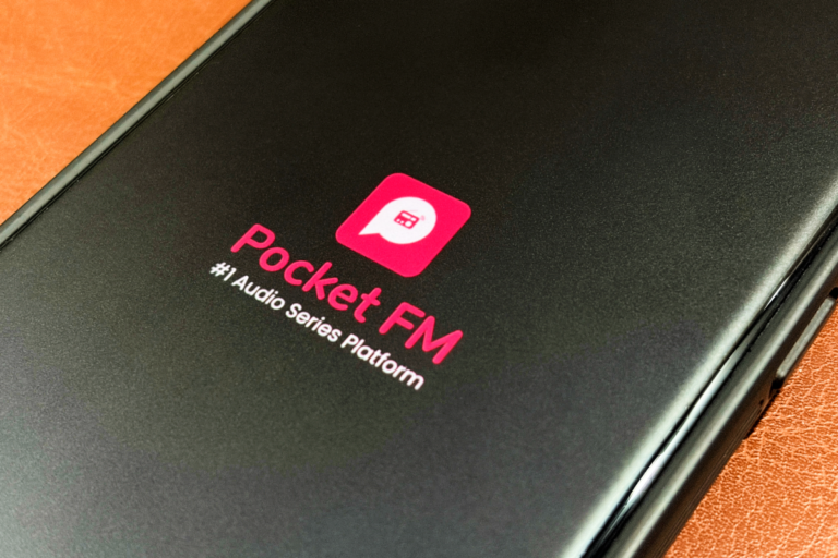 Smartphone screen displaying Pocket FM logo and text, "#1 Audio Series Platform" on a brown surface.