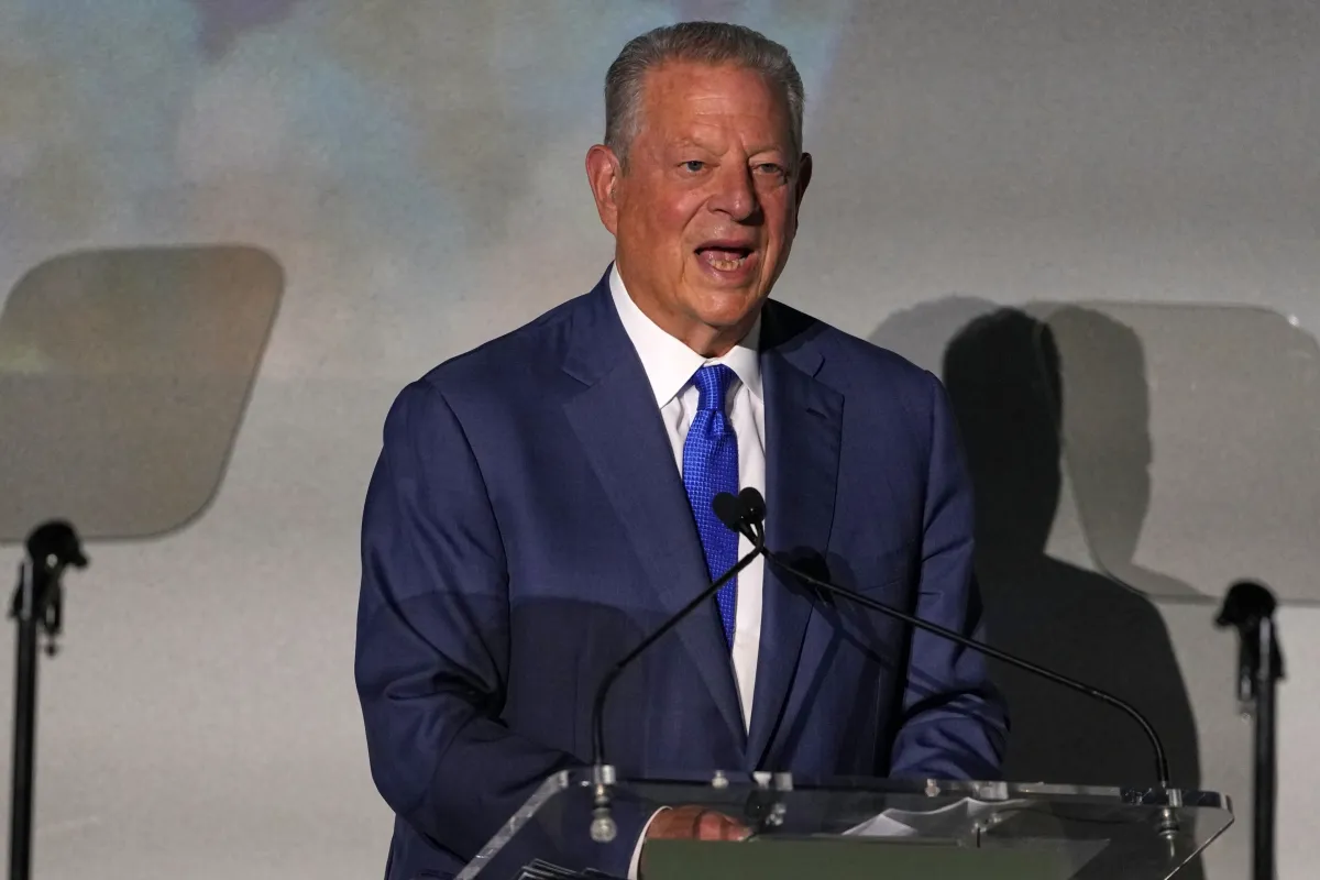 Man in a blue suit speaks at a podium with microphones, flanked by two teleprompters, against a neutral background.