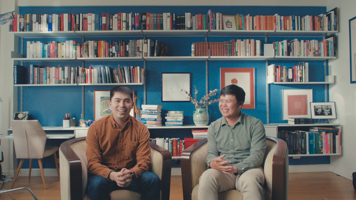 Two men seated in armchairs, smiling, with a bookshelf filled with books and framed art behind them in a warmly lit room.