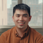 Man in brown shirt smiling in a room with large windows showing a cityscape in the background.