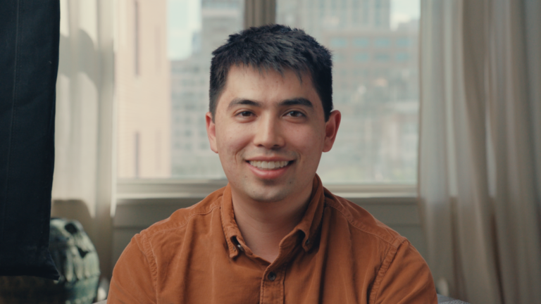 Man in brown shirt smiling in a room with large windows showing a cityscape in the background.