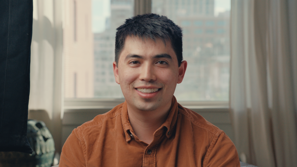 Man in brown shirt smiling in a room with large windows showing a cityscape in the background.