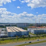 Sprawling data center complex with adjacent construction site and cranes under a partly cloudy sky, multiple cars on nearb...