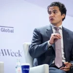 Man in a suit speaking at CERAWeek event, S&P Global logo visible, seated with bottled water and a coffee cup on a table.