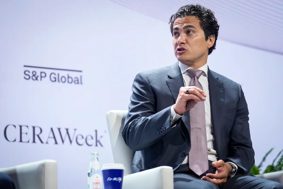 Man in a suit speaking at CERAWeek event, S&P Global logo visible, seated with bottled water and a coffee cup on a table.