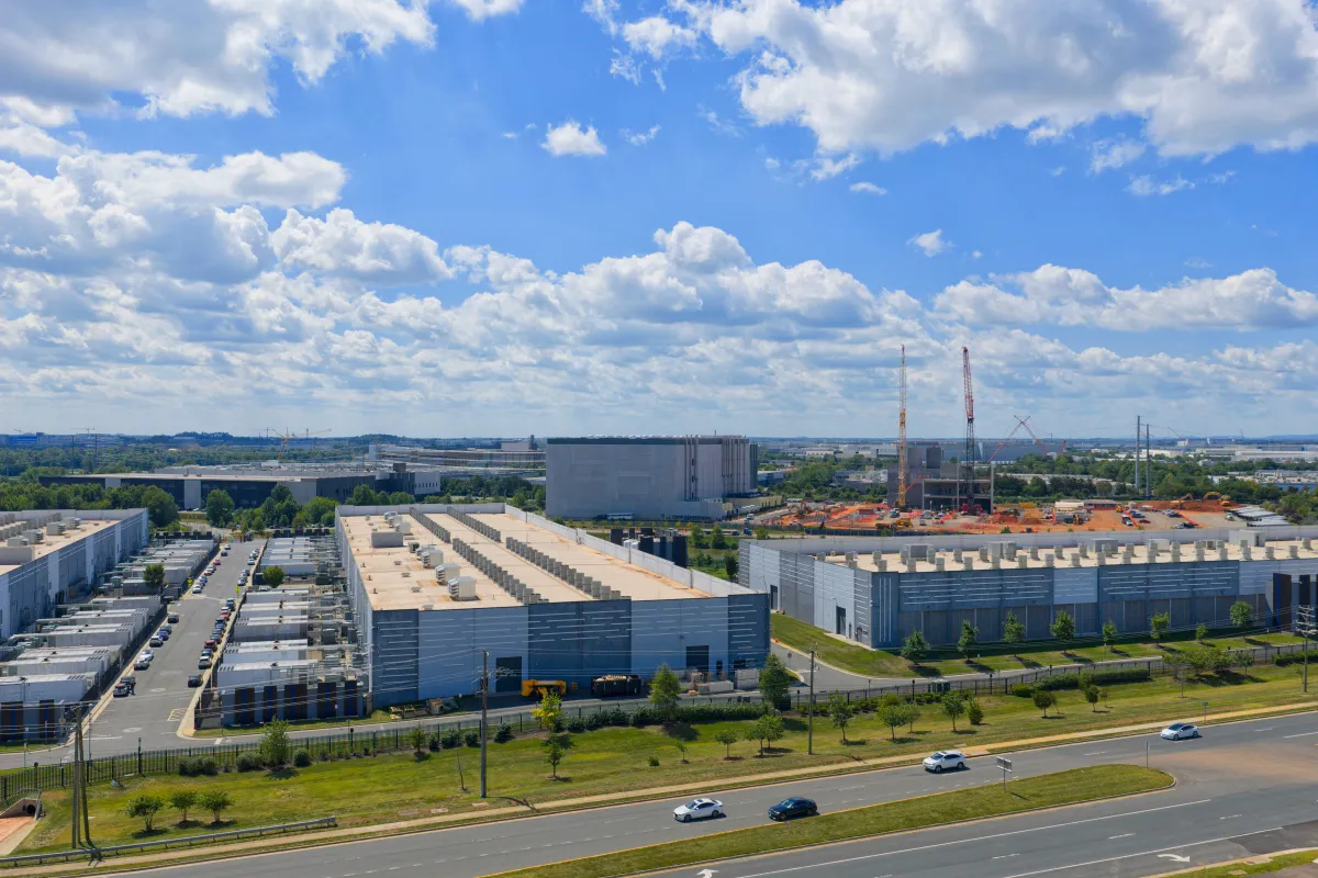Sprawling data center complex with adjacent construction site and cranes under a partly cloudy sky, multiple cars on nearb...