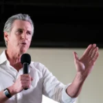 Man in a white shirt speaking into a microphone, gesturing with his hand, indoors with a neutral background.