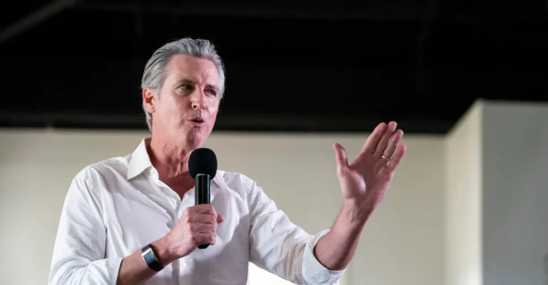 Man in a white shirt speaking into a microphone, gesturing with his hand, indoors with a neutral background.