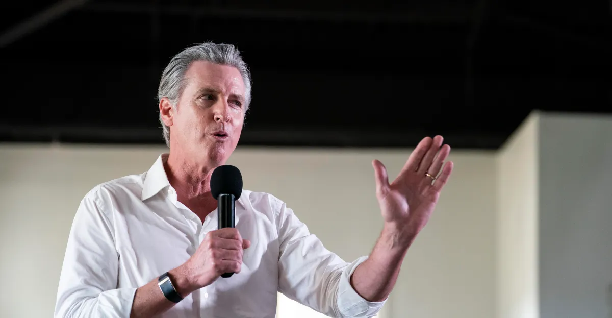 Man in a white shirt speaking into a microphone, gesturing with his hand, indoors with a neutral background.