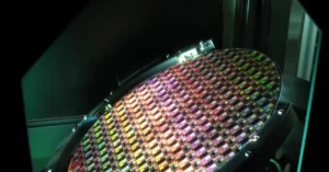 Close-up of a colorful silicon wafer with intricate microchip patterns under bright lighting in a manufacturing setting.