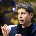 Person speaking into a microphone, gesturing with one hand, blurred background with lights.