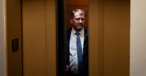 Man in a suit and tie standing inside an elevator, partially visible through closing doors, with a wall labeled 'B'.