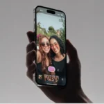 Hand holding a smartphone displaying a selfie of two smiling people with sunglasses, camera app interface visible