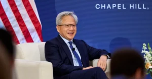 Man in a suit sitting on a stage with a microphone, American flag behind, text "Chapel Hill" partially visible on screen.
