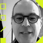 Man with glasses speaking, surrounded by yellow geometric shapes, black and white background.