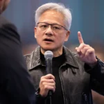 Man in a black leather jacket speaking into a microphone, gesturing with one hand, in a blurred background setting.